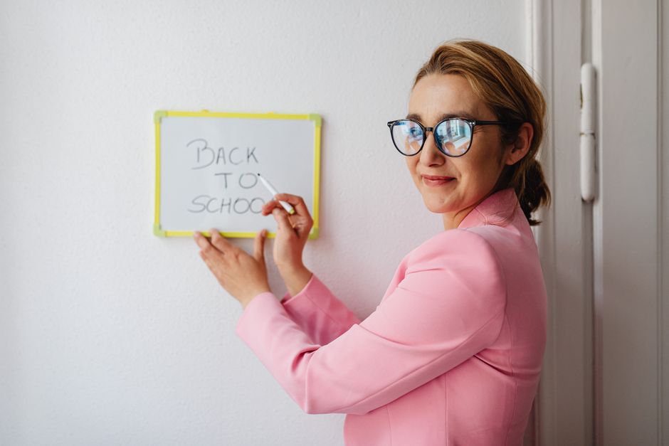 Educação Pública Brasil — A cheerful female teacher in a pink blazer writes 'Back to School' on a whiteboard.
